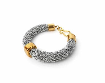 Puede incluir: Una pulsera de malla plateada con detalles dorados. La pulsera presenta un diseño tejido y se asegura con un cierre dorado. Cuenta con extremos cuadrados dorados.