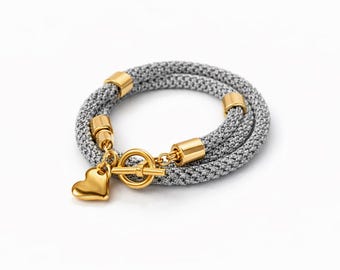 Puede incluir: Una pulsera envolvente de malla plateada con detalles dorados y un dije de corazón. La pulsera tiene un cierre de palanca y se muestra sobre un fondo blanco. Una pieza de joyería elegante.