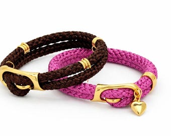 Puede incluir: Dos pulseras de cuerda. Una es marrón con herrajes dorados. La otra es rosa con herrajes dorados y un dije de corazón. Ambas pulseras tienen un diseño de cuerda trenzada.