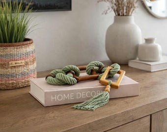 Puede incluir: Una llave decorativa de madera con detalles de cuerda verde y una borla descansa sobre un libro que dice "HOME DECOR". La llave está colocada sobre una superficie de madera, con un jarrón y una pintura de paisaje enmarcada en el fondo.