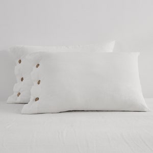 May include: Two white pillowcases with a textured appearance. Each pillowcase has a row of wooden buttons along the side. The pillowcases are rectangular and rest on a white surface.