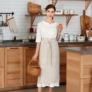 May include: A woman wearing a white linen apron with a brown linen skirt and a white shirt. She is holding a wicker basket and a small orange. The apron has a tie at the waist and a pocket on the front. The woman is standing in a kitchen with wooden cabinets and white tiled walls.