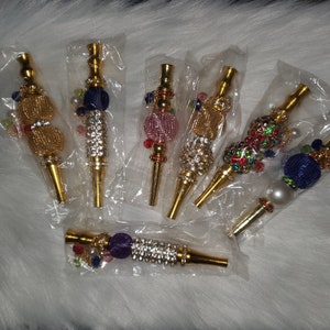 May include: Six gold-toned smoking pipes with decorative embellishments. Each pipe features a different color scheme with rhinestones and beads. The pipes are packaged individually in clear plastic bags.