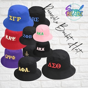 May include: A collection of reversible bucket hats featuring various Greek letter designs, including AKA, Delta Sigma Theta, Kappa Alpha Psi, Omega Psi Phi, Sigma Gamma Rho, Zeta Phi Beta, and Alpha Phi Alpha. The hats are black, red, blue, purple, and pink.