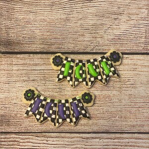 May include: Two black and white checkered cookies decorated with green and purple icing. The cookies are shaped like banners and spell out the word "FOUR".