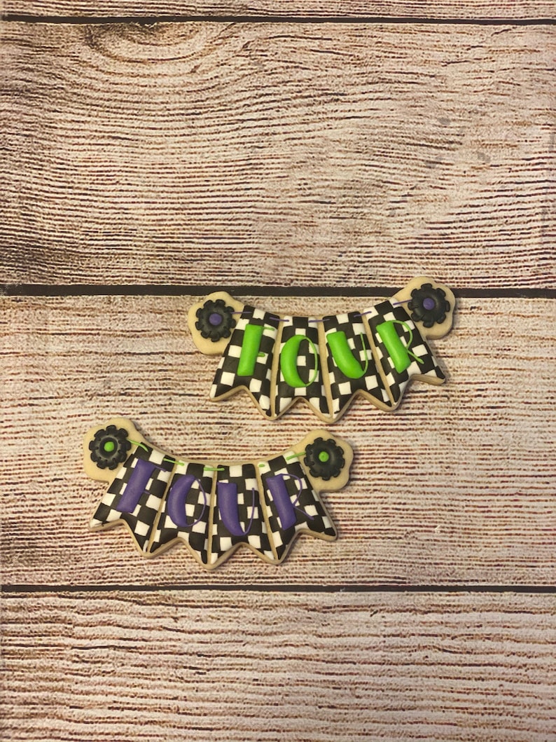 May include: Two black and white checkered cookies decorated with green and purple icing. The cookies are shaped like banners and spell out the word "FOUR".