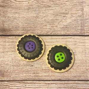 May include: Two decorated sugar cookies shaped like tyre rims. One cookie has a purple centre with black dots and the other has a green centre with black dots.