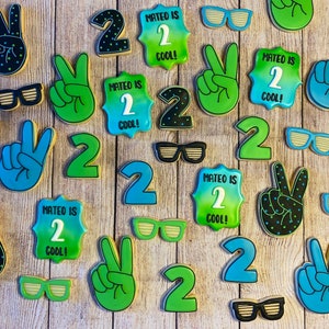 May include: A collection of decorated sugar cookies with various designs, including the number "2", peace signs, and sunglasses. The cookies are decorated in shades of green, blue, and black with white icing. The text "Mateo is 2 Cool!" is written on several of the cookies.