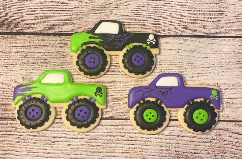 May include: Three monster truck cookies decorated with green, purple and black icing. Each cookie has a skull and crossbones design and a lightning bolt pattern.