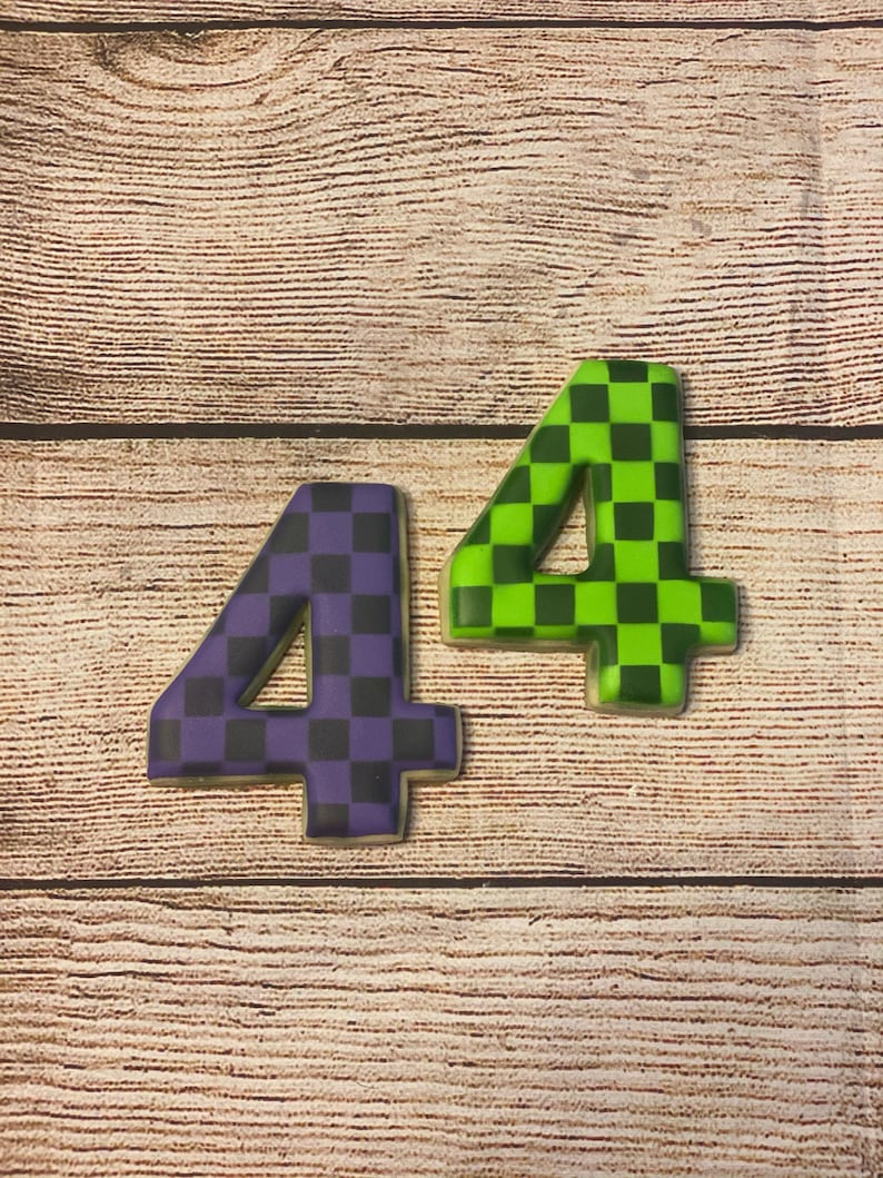 May include: Two cookies shaped like the number four, decorated with a checkered pattern. One cookie is purple and black, the other is green and black.