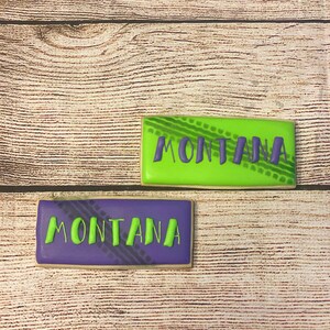 May include: Two decorated sugar cookies with the word "MONTANA" written in green icing on a purple cookie and green icing on a green cookie. The cookies have a tyre tread pattern on them.