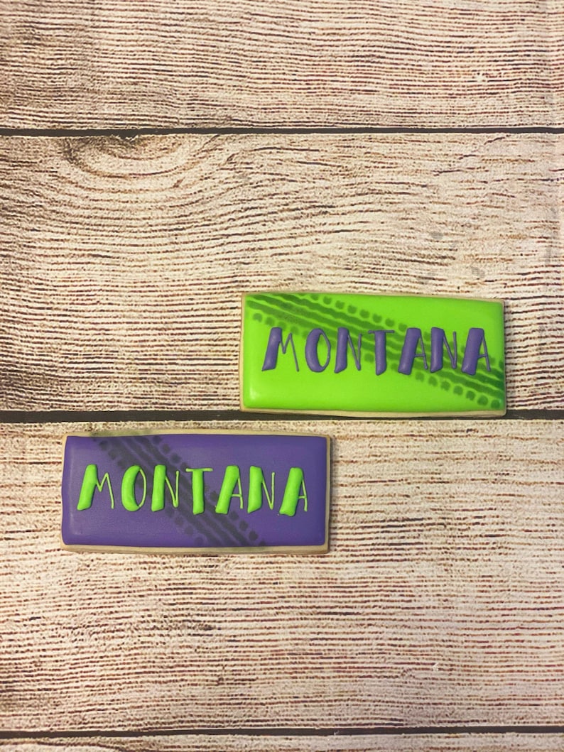 May include: Two decorated sugar cookies with the word "MONTANA" written in green icing on a purple cookie and green icing on a green cookie. The cookies have a tyre tread pattern on them.