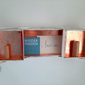 May include: A white wooden shelf with three compartments, each with a copper interior. The middle compartment has a blue card with the text "WIEDER FINDEN" and a white card with the text "i love us". The other two compartments have wooden blocks.