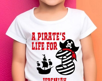 Può includere: Una maglietta bianca con la scritta "A Pirate's Life For" e il nome "Jeremiah" in rosso. La maglietta presenta una nave pirata e il numero 3 con un cappello da pirata. Il bambino indossa occhiali da sole blu e pantaloncini a righe colorate.
