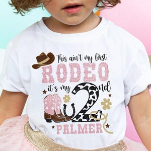 May include: White t-shirt with a rodeo design. The shirt has the text "This ain't my first RODEO it's my 2nd" with a cowboy hat, boots, horseshoe, and the name "PALMER". The outfit includes a pink tutu skirt.