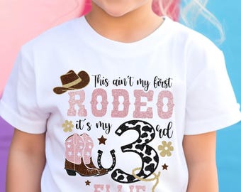 May include: A white short-sleeved t-shirt featuring a graphic print with a brown cowboy hat, pink glittery cowboy boots, a horseshoe, and the text: This ain't my first RODEO it's my 3rd ELLIE.