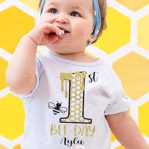 Puede incluir: Un body blanco con un número uno de purpurina dorada y un diseño de miel que gotea. El body también tiene una abeja negra y el texto "Bee-Day Ayla 1st".