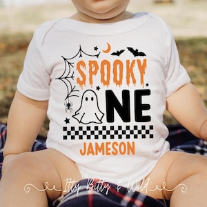 May include: A white short-sleeved baby bodysuit featuring a graphic with the orange text SPOOKY and black text ONE. The design includes a ghost, a spider web, a spider, bats, a crescent moon, stars, and a black and white checkered pattern.