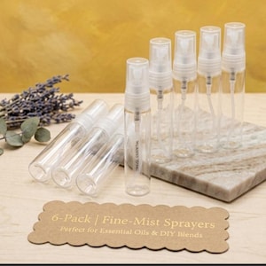 May include: A set of six clear fine-mist spray bottles with white spray tops. The bottles are arranged on a marble-patterned block. A sprig of lavender and a brown tag with text are also in the image. The text on the tag reads "6-Pack | Fine-Mist Sprayers".