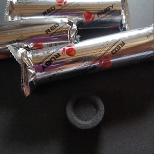 Puede incluir: Varios paquetes envueltos en papel de aluminio plateado de carbón de encendido rápido de la marca Ruby, con detalles en rojo y naranja, y un solo disco de carbón. Los paquetes tienen el texto "RED" y "RUBY" impreso.
