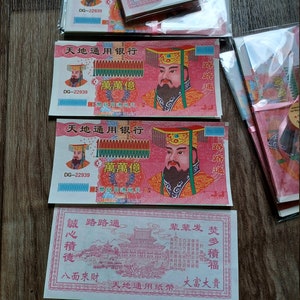 May include: Several stacks of colorful paper money with Chinese characters and illustrations of a man in a hat. The bills are predominantly red, green, and gold, with intricate designs and text. Some bills are in clear plastic packaging.