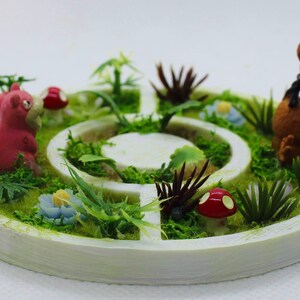 Peut inclure: Une scène de jardin miniature avec un personnage Pokémon rose et blanc, un personnage Pokémon brun et une variété de plantes vertes et de champignons rouges. La scène est installée dans un pot rond blanc avec une base de mousse verte.