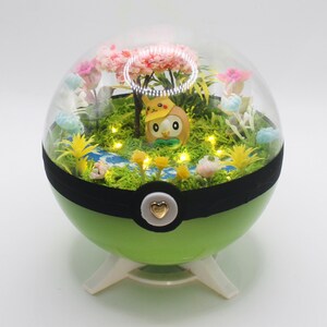 Könnte beinhalten: Ein kugelförmiges grünes Terrarium mit einer transparenten Kuppel auf einem weißen Ständer. Im Inneren befindet sich eine kleine braune eulenartige Figur mit einem gelben Hut. Die Szene enthält grünes Moos, einen blauen Bach, rosa Blüten und LED-Lichter.