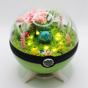 Op de afbeelding: Een bolvormig groen terrarium met een Bulbasaur-figuur, een miniatuur roze kersenbloesemboom en kleine kunstplanten op groen mos. Het display wordt verlicht door interne LED-lampjes en staat op een witte standaard.