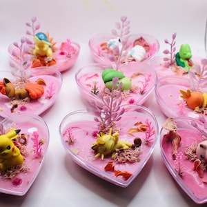 Peut inclure: Plusieurs récipients en forme de cœur remplis de liquide rose et de figurines miniatures. Chaque récipient présente un personnage Pokémon différent, entouré d'éléments décoratifs tels que des plantes artificielles et de la mousse. L'ensemble est mignon et fantaisiste.