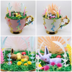 Peut inclure: Une tasse à thé rose avec un intérieur blanc et une bordure dorée est remplie de mousse verte, de fleurs colorées et de petites figurines de personnages Pokémon. La tasse à thé est décorée de feuilles vertes et repose sur une surface blanche.