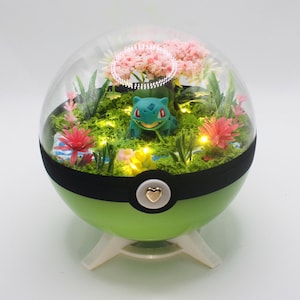 Puede incluir: Un terrario esférico verde con una figura de Bulbasaur azul claro sobre musgo verde, árboles de cerezo en flor rosas, plantas pequeñas coloridas y luces LED cálidas integradas, todo dentro de una cúpula transparente con un botón de corazón.