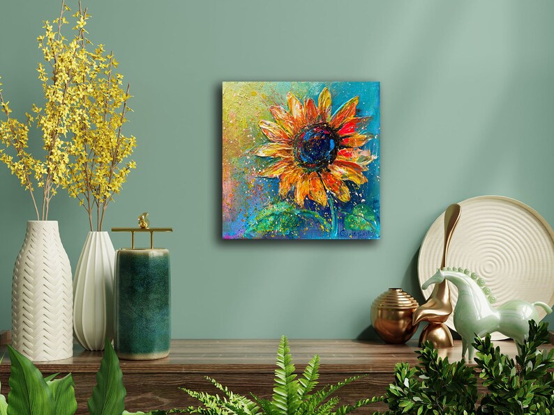 May include: A vibrant painting of a sunflower with orange and yellow petals and a dark blue center. The artwork is on a square canvas, with a textured, impressionistic style. The background is a mix of blue, green, and yellow.