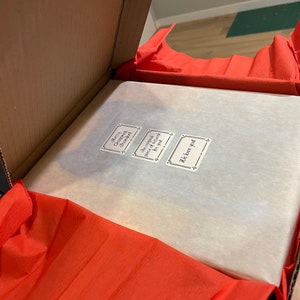 May include: An open cardboard box with red tissue paper and a wrapped package inside. Three small stickers are visible on the package, with text that reads "Merry Christmas, Sweetheart", "the original piece of artwork for you", and "We love you".