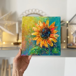 May include: Square canvas painting of a sunflower. The sunflower has yellow and orange petals with a dark blue center. The background is a mix of blue and yellow with green leaves at the bottom. The painting has a textured, impressionistic style.