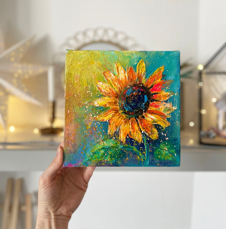 May include: Square canvas painting of a sunflower. The sunflower has yellow and orange petals with a dark blue center. The background is a mix of blue and yellow with green leaves at the bottom. The painting has a textured, impressionistic style.