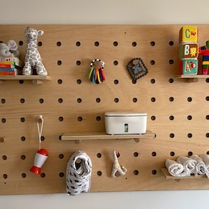May include: A wooden pegboard with various baby items. Includes stuffed animals, books, a white container, toys, and rolled-up washcloths. The pegboard has multiple holes for attaching shelves and hanging items.
