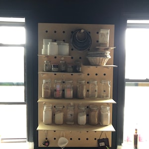 May include: A wooden pegboard with shelves displays various kitchen items. Clear glass jars filled with spices and powders are arranged on the shelves. White ceramic containers, metal ring molds, and a woven basket are also visible, suggesting a baking or cooking theme.