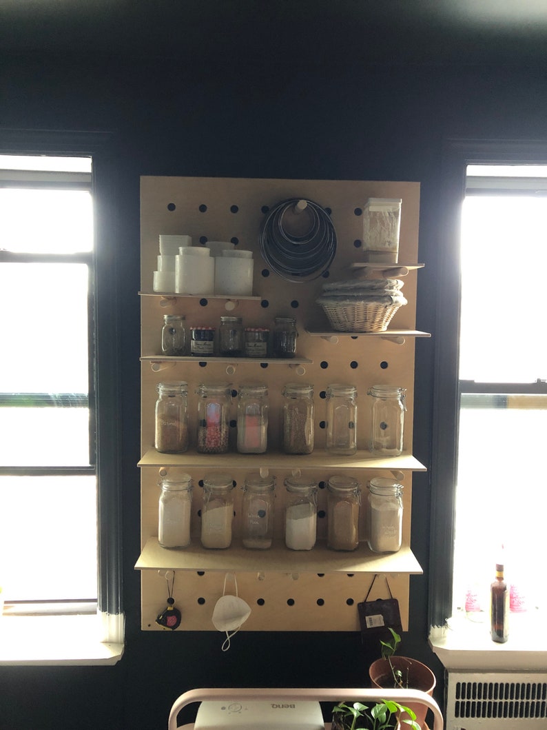 May include: A wooden pegboard with shelves displays various kitchen items. Clear glass jars filled with spices and powders are arranged on the shelves. White ceramic containers, metal ring molds, and a woven basket are also visible, suggesting a baking or cooking theme.
