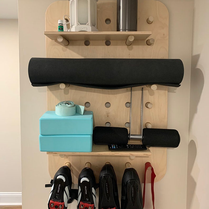 May include: A wooden wall-mounted organizer with a variety of fitness equipment. It includes a rolled-up black yoga mat, blue yoga blocks, a resistance band, and cycling shoes. A small shelf holds a lantern and a water bottle.