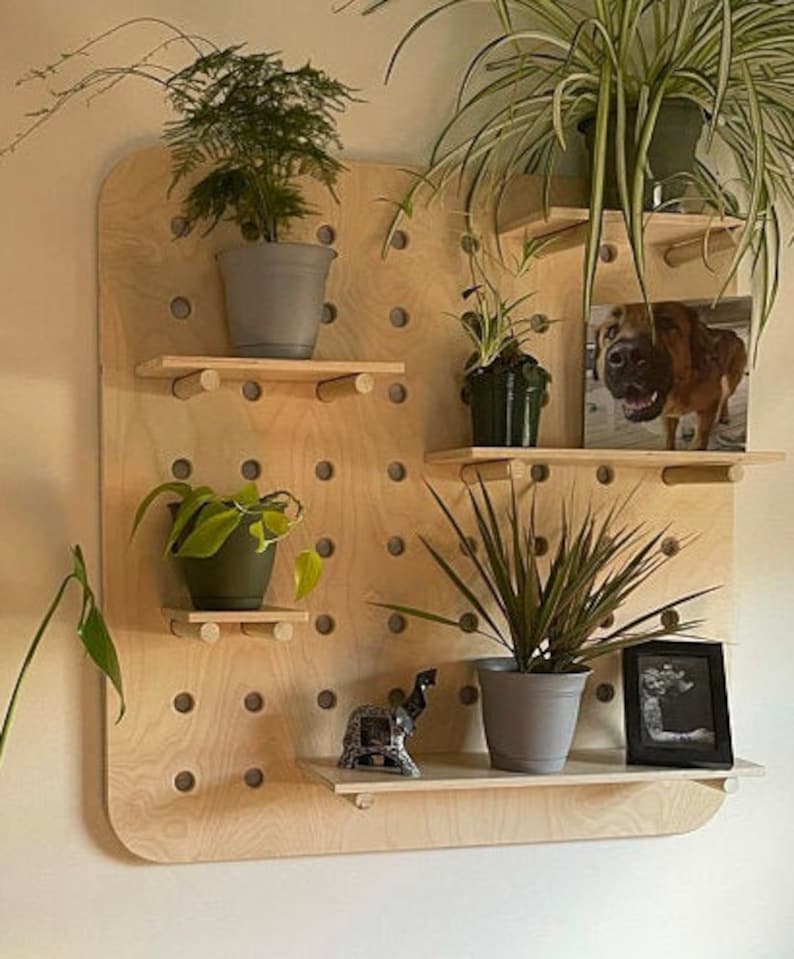 May include: A wooden pegboard with multiple shelves, displaying potted plants, a framed photo of a dog, and a small decorative elephant. The pegboard is made of light-colored wood and has a grid of evenly spaced holes.