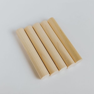 May include: Five wooden dowels, all the same size, are arranged in a semi-circle on a white surface. The dowels are light brown in color and have a smooth, rounded finish.