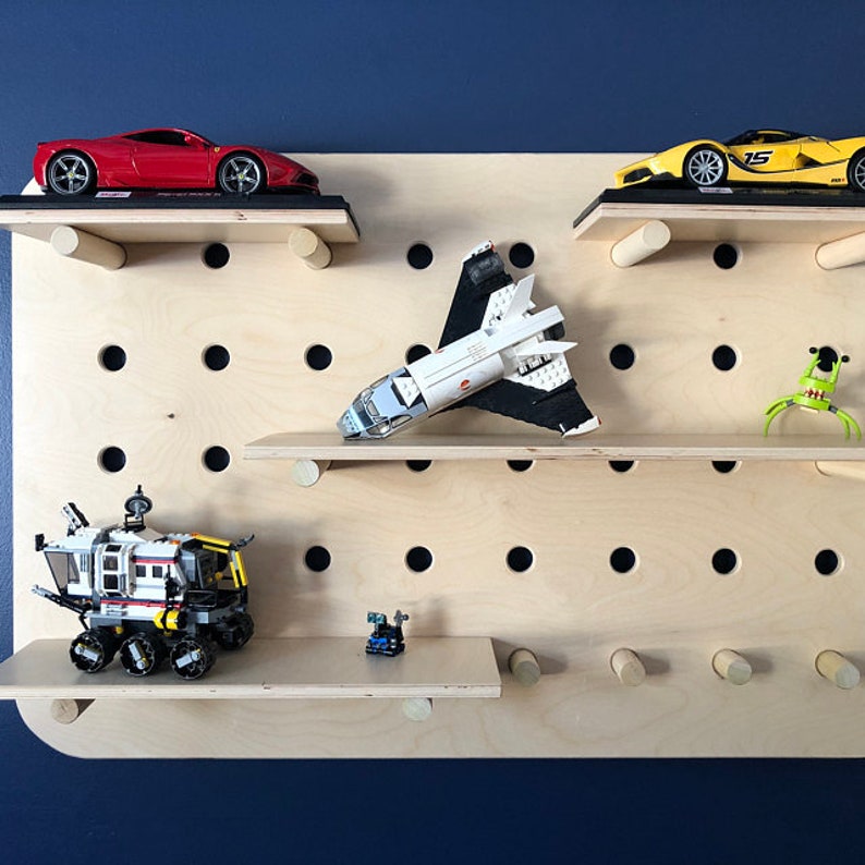 May include: A wooden pegboard with multiple shelves displays toy cars, a space shuttle, and other small toys. The pegboard is mounted on a dark blue wall. The toys are in various colors, including red, yellow, white, and black.