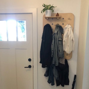 May include: A wooden wall-mounted coat rack with a small shelf holding a potted plant and a small statue. The rack has several pegs holding a black coat, a denim jacket, and a white jacket. An umbrella leans against the wall.