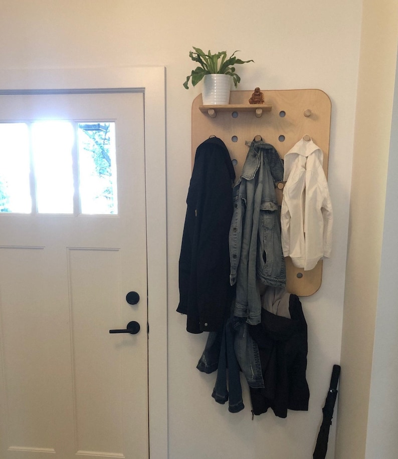 May include: A wooden wall-mounted coat rack with a small shelf holding a potted plant and a small statue. The rack has several pegs holding a black coat, a denim jacket, and a white jacket. An umbrella leans against the wall.