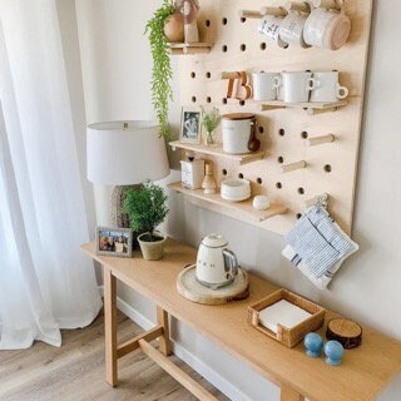 May include: A light wood bench with a lamp, small potted plants, and a pegboard shelf. The pegboard holds mugs, small shelves, and decorative items. A white teapot sits on a wooden tray on the bench.