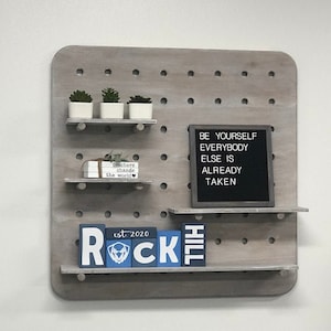 May include: A gray pegboard with three small shelves. The top shelf holds three small potted succulents. The second shelf has a stack of books. The third shelf has wooden blocks that spell "ROCK HILL". A framed sign reads "BE YOURSELF EVERYBODY ELSE IS ALREADY TAKEN."