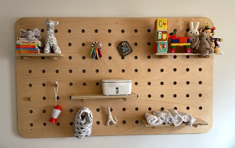 May include: A wooden pegboard with various baby items. Includes stuffed animals, books, a white container, toys, and rolled-up washcloths. The pegboard has multiple holes for attaching shelves and hanging items.