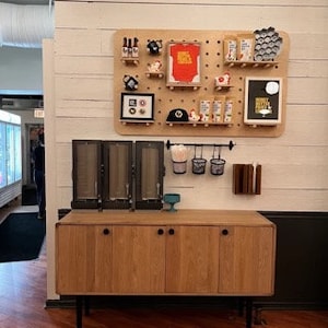 May include: A wooden cabinet with four doors and black legs. Above it, a pegboard displays various items, including small bottles, a framed picture, and a baseball cap. The wall is partially covered with white wooden planks.