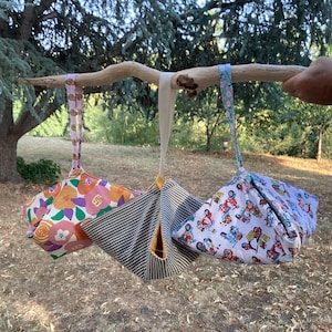 Può includere: Tre borse in tessuto con cinturini da polso appese a un ramo di legno. La borsa a sinistra presenta un motivo floreale colorato, quella centrale ha righe bianche e nere con interno giallo e quella a destra mostra un motivo con piccoli scooter.