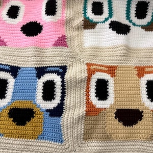 May include: A crochet blanket featuring four square panels, each displaying a different stylized animal face. The faces are rendered in pink, white, blue, and tan yarn with black accents, all framed by a neutral beige border connecting the squares.